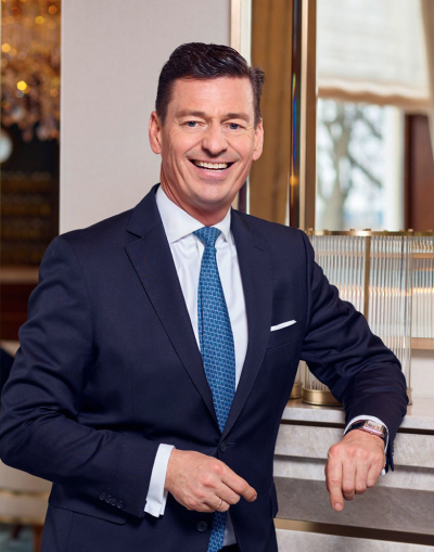 Ein Mann in einem dunklen Anzug und mit blauer Krawatte steht in einem Haus, lächelt selbstbewusst und stützt sich mit einer Hand auf eine Tischplatte. Im Hintergrund sind ein Fenster und eine dekorative Beleuchtung zu sehen.