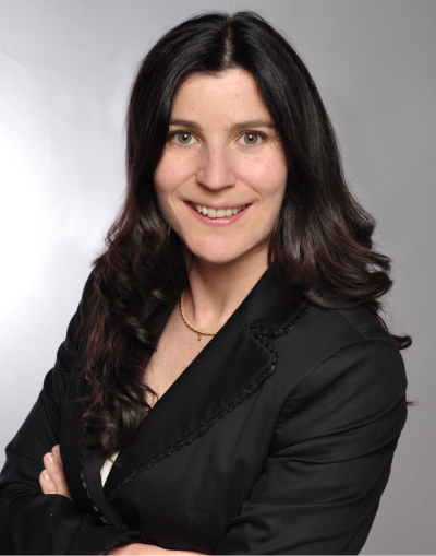 Eine Frau mit langen, dunklen, gewellten Haaren, die einen schwarzen Blazer und eine goldene Halskette trägt, lächelt in die Kamera vor einem schlichten grauen Hintergrund. Ihre Arme sind verschränkt.