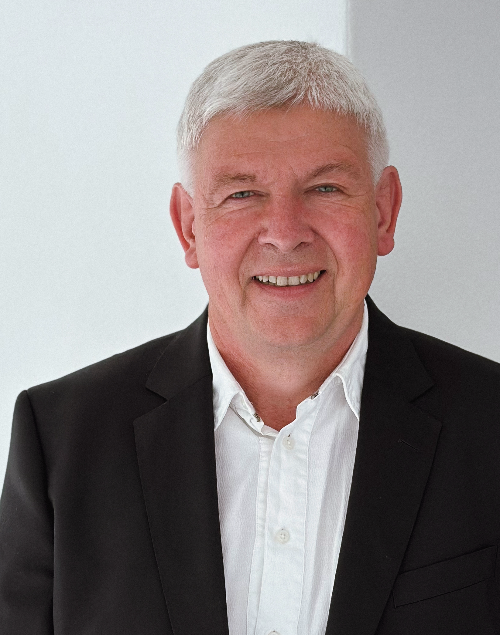 thuy Ein lächelnder älterer Mann mit kurzen grauen Haaren, der einen schwarzen Blazer über einem weißen Button-up-Hemd trägt, steht vor einem schlichten weißen Hintergrund.