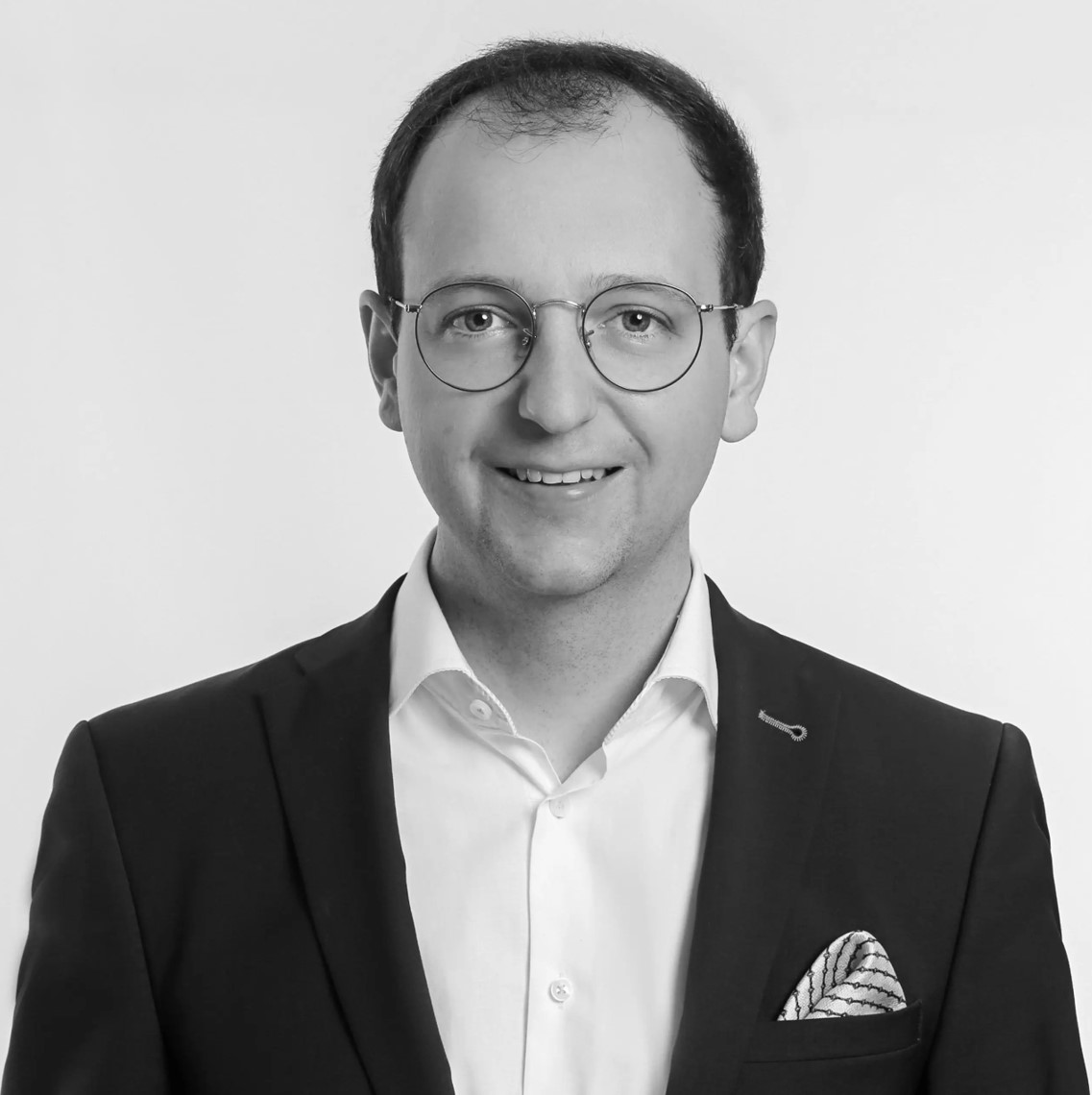 Ein Mann, der eine Brille und eine Anzugsjacke mit Einstecktuch trägt, lächelt in die Kamera. Er hat kurzes Haar und steht vor einem schlichten, hellen Hintergrund. Das Bild ist in schwarz-weiß gehalten.