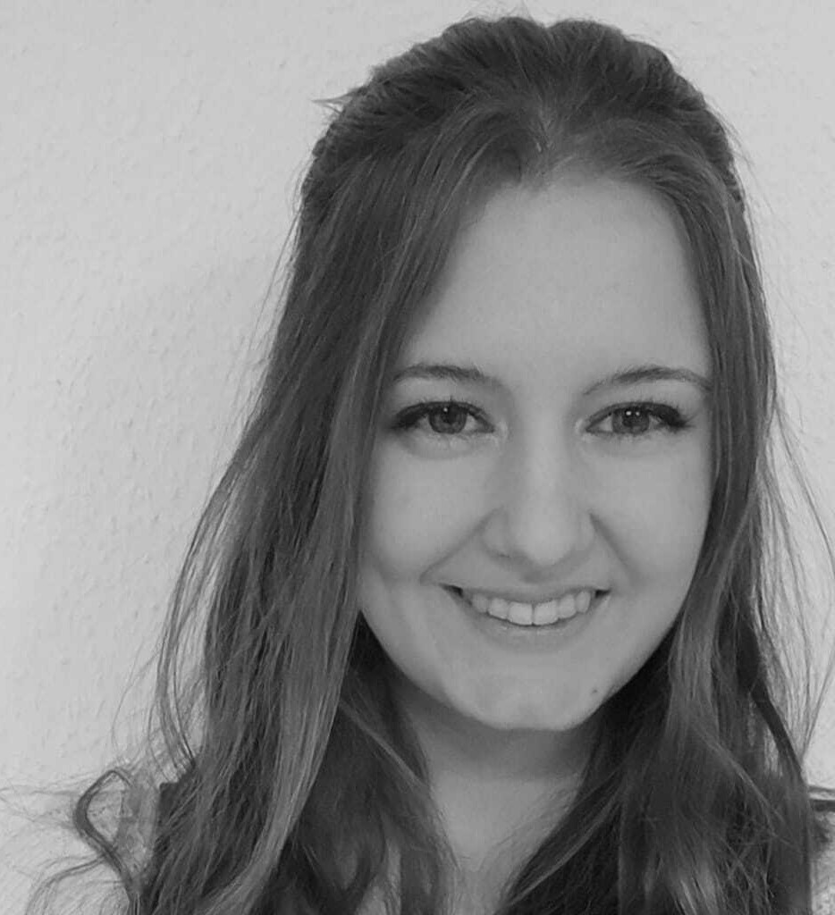 Schwarz-Weiß-Foto einer jungen Frau mit langen Haaren, die in die Kamera lächelt, vor einem schlichten hellen Hintergrund.