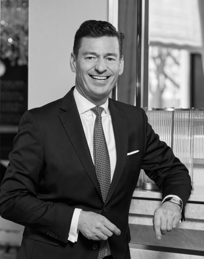 Ein Mann in Anzug und Krawatte lächelt, während er in einem Haus steht und einen Arm auf einen Tresen stützt. Er hat kurzes dunkles Haar und trägt eine Uhr. Das Bild ist schwarz-weiß.