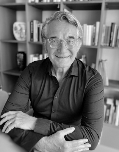 Ein lächelnder älterer Mann mit Brille und grauem Haar sitzt an einem Tisch mit verschränkten Armen vor einem mit Büchern und Dekorationsgegenständen gefüllten Bücherregal. Das Foto ist in schwarz-weiß gehalten.