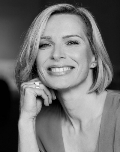 Schwarz-Weiß-Foto einer lächelnden Frau mit kurzen, blonden Haaren, die ein helles Oberteil trägt. Sie stützt ihr Kinn auf ihre Hand und blickt selbstbewusst in die Kamera.