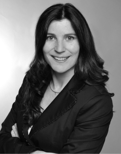 Eine Frau mit langen, gewellten dunklen Haaren und einem dunklen Blazer lächelt mit verschränkten Armen in die Kamera, vor einem schlichten, hellen Hintergrund. Das Bild ist in schwarz-weiß gehalten.