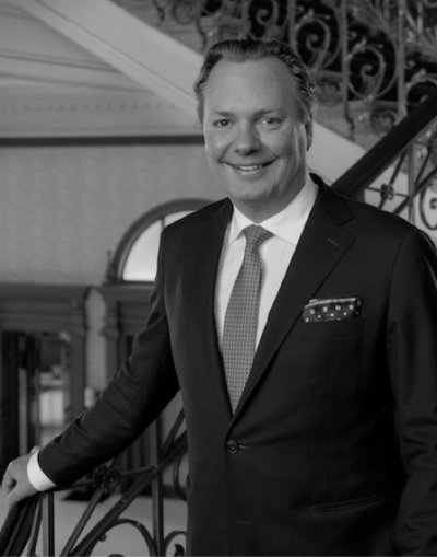 Ein Mann in Anzug und Krawatte steht lächelnd an einer verschnörkelten Treppe, eine Hand auf das Geländer gestützt, in einer eleganten Inneneinrichtung.