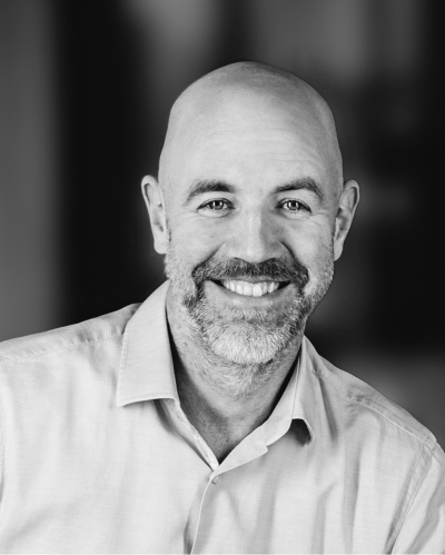 Schwarz-Weiß-Porträt eines Mannes mittleren Alters mit Glatze und Bart, der freundlich in die Kamera lächelt. Er trägt ein helles Hemd mit Kragen, der Hintergrund ist unscharf.
