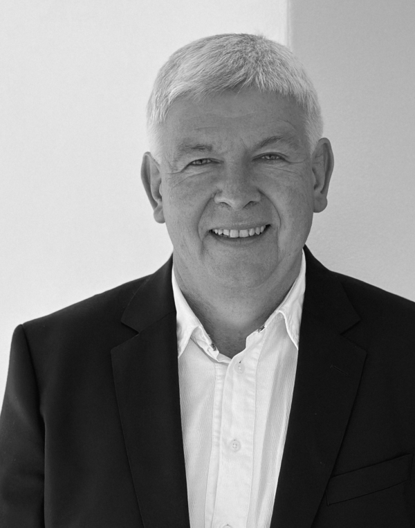 Ein lächelnder älterer Mann mit kurzen Haaren, der einen dunklen Blazer über einem weißen Button-up-Hemd trägt, steht vor einem schlichten hellen Hintergrund.