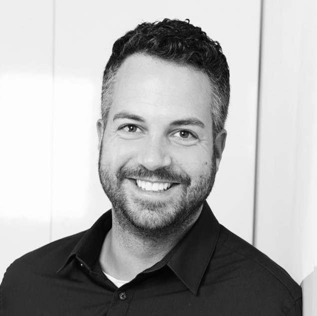 Ein Mann mit kurzen lockigen Haaren und einem Bart lächelt in die Kamera. Er trägt ein dunkles Hemd und steht vor einem hellen Hintergrund. Das Foto ist in schwarz-weiß gehalten.